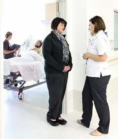 A nurse standing at the bedside of a male patient with two women talking in a nearby hallway/